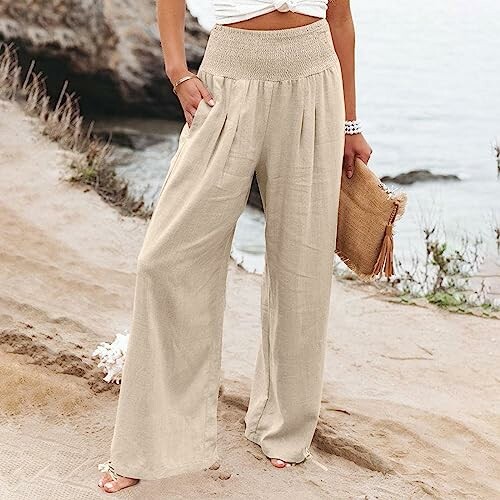 Woman in beige wide-leg pants holding a woven clutch by the beach.