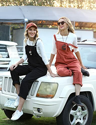 Two women sitting on a Jeep wearing casual overalls and sunglasses.